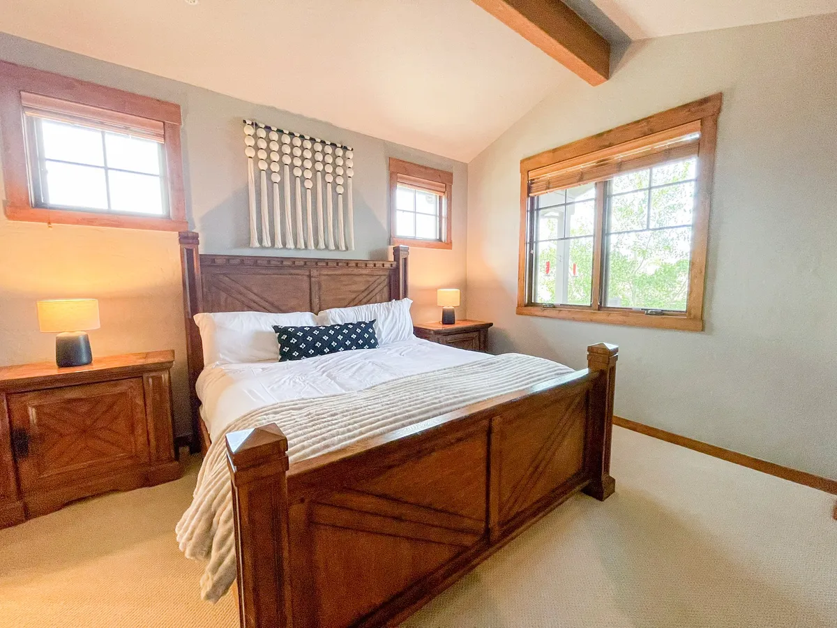 A tranquil bedroom featuring a large bed adorned with decorative pillows, wooden furniture, and windows that let in ample natural light. The space exudes comfort and invites restful sleep or relaxation during the day.