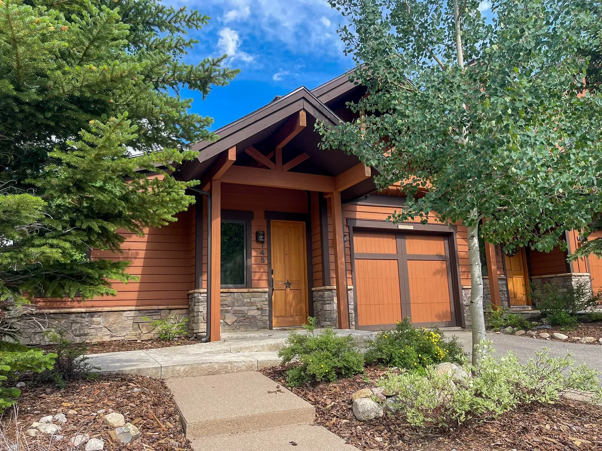 The image captures the villa’s inviting stone façade set amongst aspen trees and lush landscaping, with a welcoming front door that hints at a cozy homecoming. The scene suggests an approachable, family-friendly atmosphere in a scenic mountain setting.