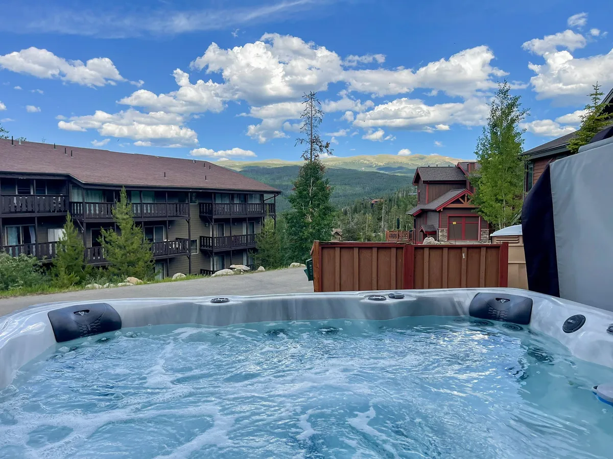 A tranquil exterior scene featuring a private hot tub surrounded by trees, with sweeping mountain views in the background. People are relaxing in the hot tub, soaking up the natural beauty and enjoying a moment of calm.
