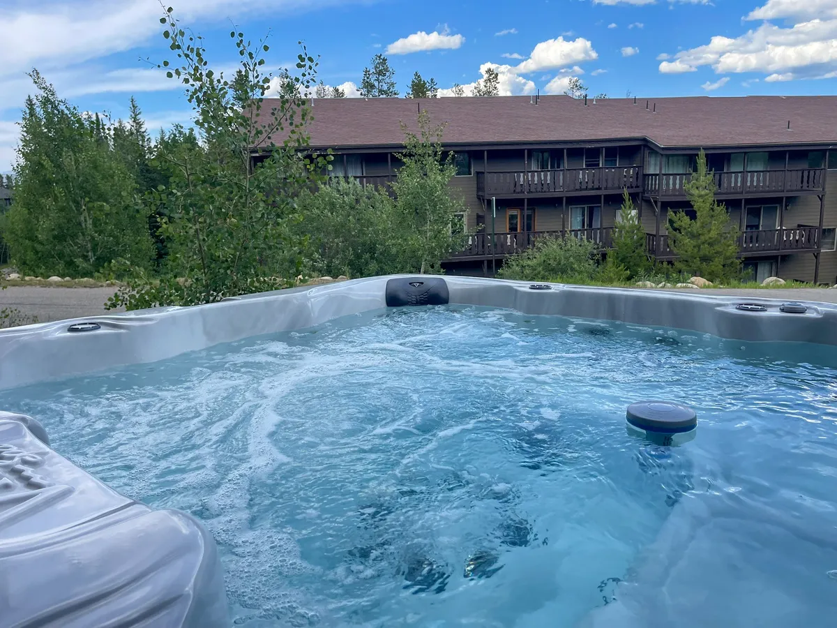 A hot tub nestled among trees with a building in the background invites relaxation. The setting is tranquil, offering a perfect spot to unwind after a day of adventure, with options for stargazing or socializing in the evening.