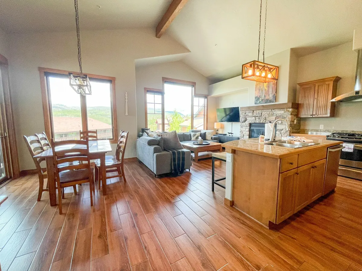 An open-plan interior view highlighting a large dining table set for a family meal, cozy sofa, kitchen island, fireplace, and expansive windows letting in natural light. The setting is warm and welcoming, emphasizing communal dining and relaxation.