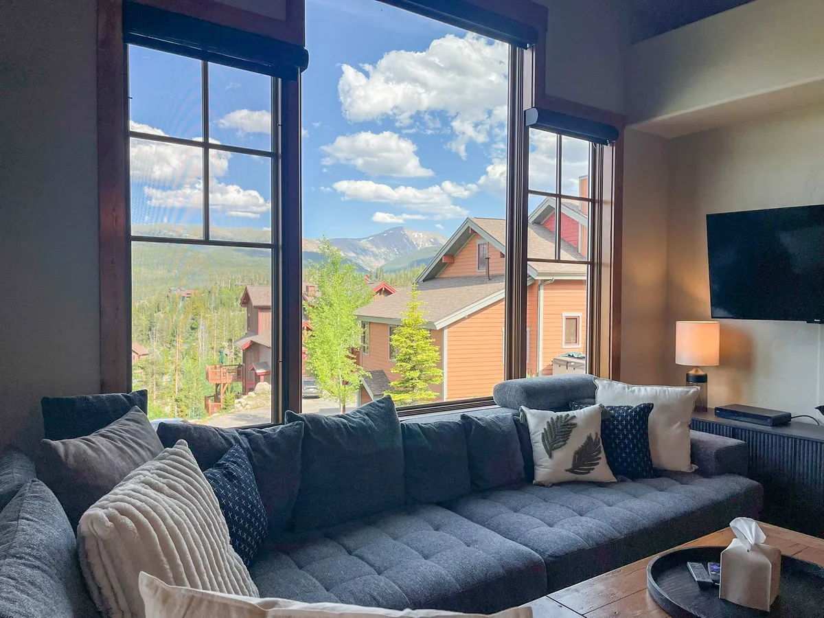 A welcoming living space with a comfortable sectional sofa, decorative pillows with natural motifs, and a wall-mounted TV. Floor-to-ceiling windows frame spectacular mountains, and the arrangement invites both casual lounging and group activities like board games or movie nights.