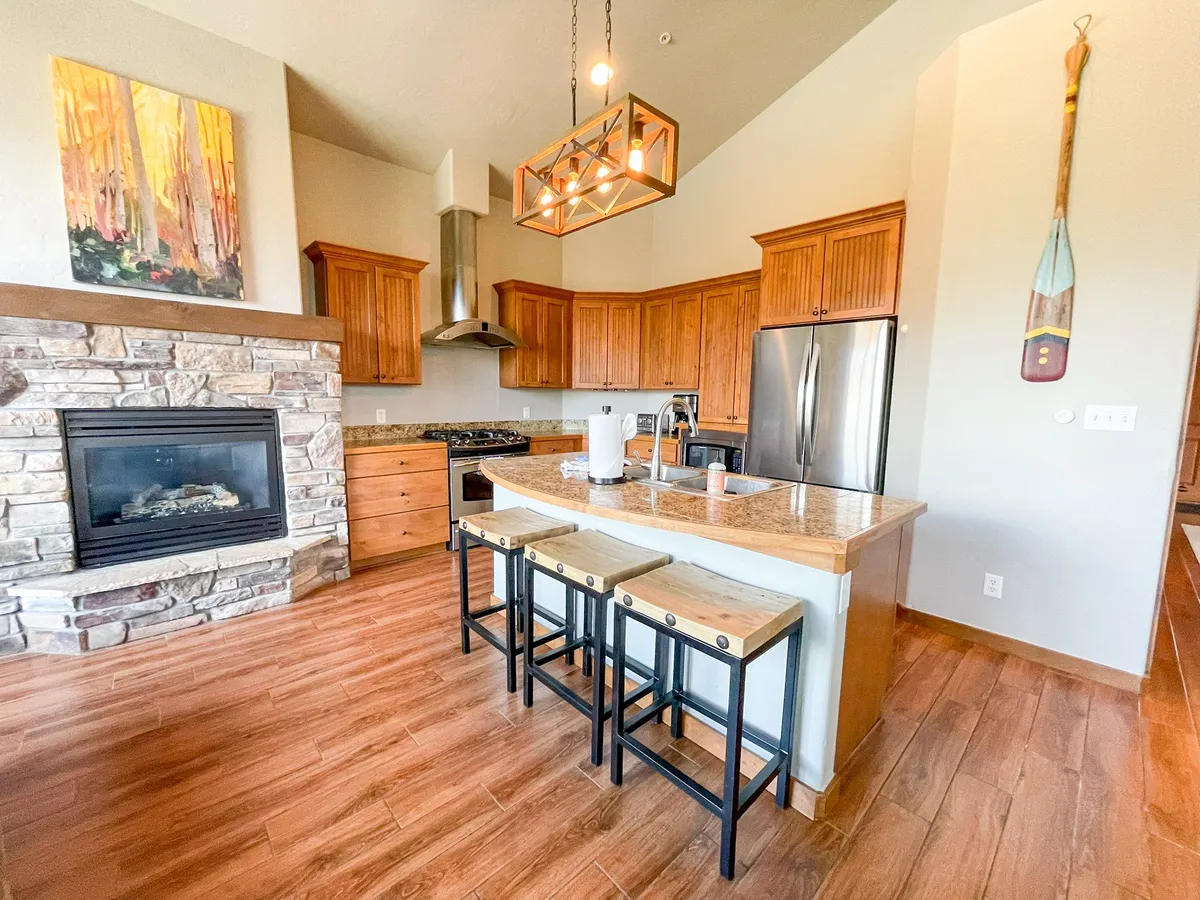 A view of the kitchen island in the foreground with a rustic stone fireplace behind, complemented by wooden cabinetry and colorful artwork. The scene suggests intimate family gatherings, coffee breaks, and storytelling in a cozy, inviting environment.