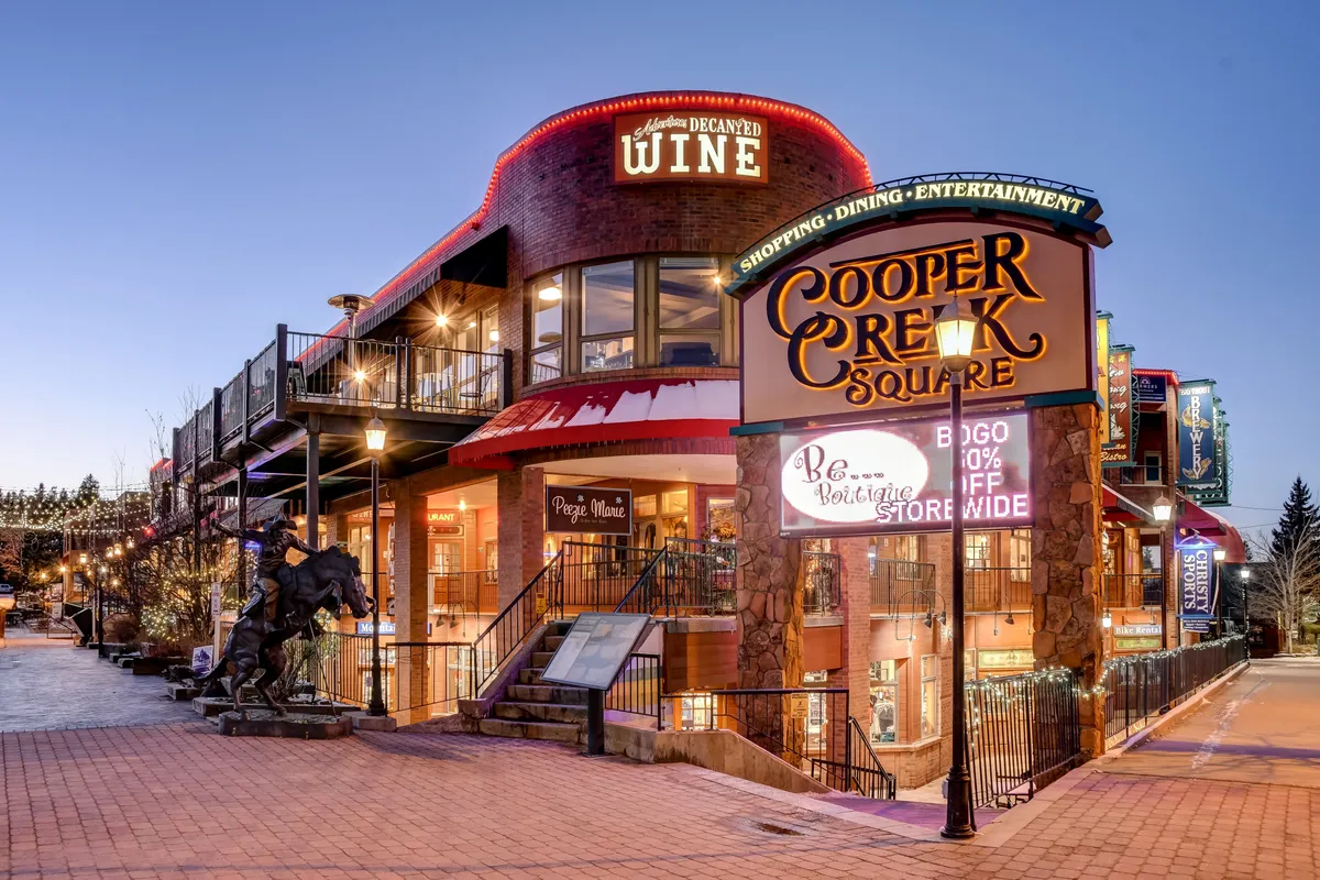 An inviting street scene featuring the Cooper Creek Square sign, a wine shop, outdoor seating, and decorative lights. The area is bustling with energy, suggesting lively shopping, dining, and community events in the heart of downtown.