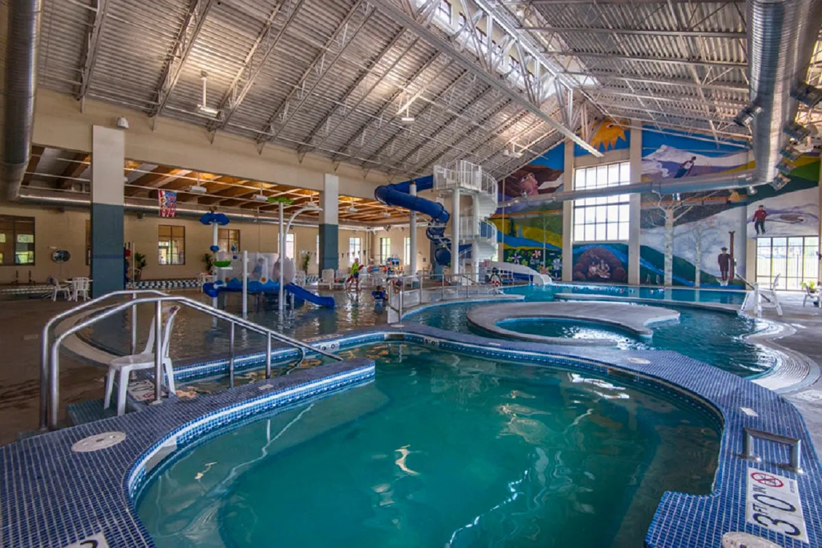 A lively, colorful interior view of the Grand Park Rec Center's swimming pool area, complete with a water slide and children playing. The space exudes energy, fun, and family-friendly activity.