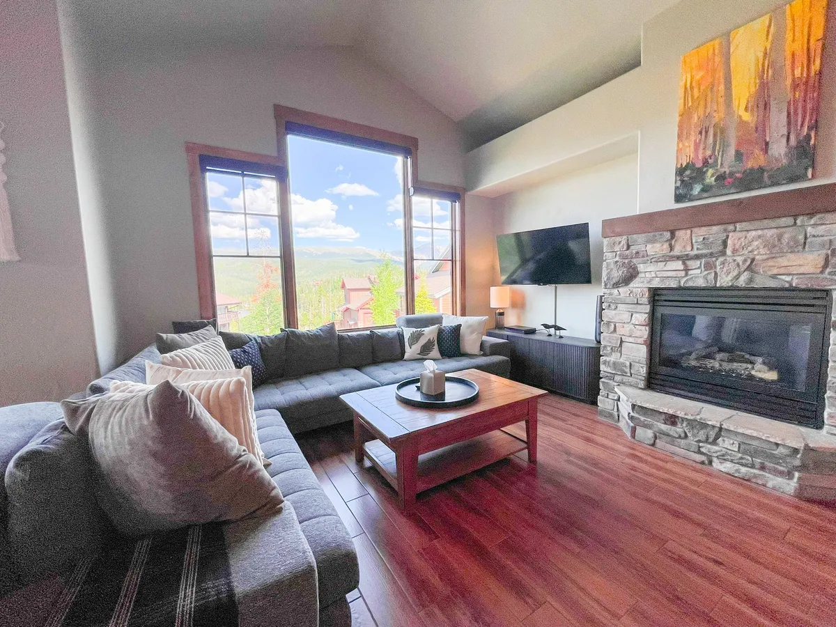 This image features a large sectional sofa arranged around a striking stone fireplace with a wall-mounted HD Smart TV above. Expansive windows reveal breathtaking mountain views, and a coffee table anchors the gathering space, evoking warmth and comfort. The layout is clearly designed for group relaxation, entertainment, and soaking up the scenery.