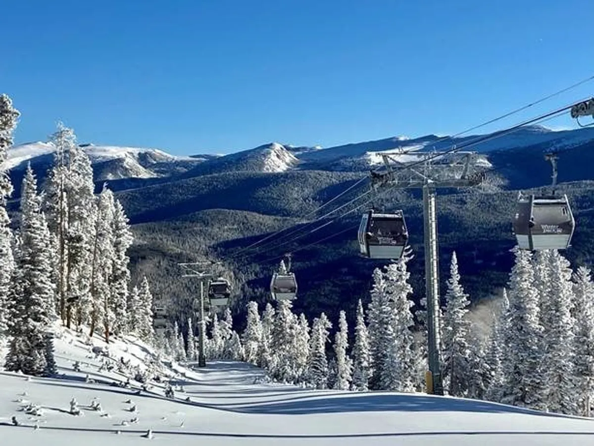 A ski lift glides above snow-covered trees with a stunning mountain range in the background and a clear blue sky overhead. Skiers and snowboarders can be seen enjoying the slopes, embodying the thrill and beauty of a classic ski resort experience.